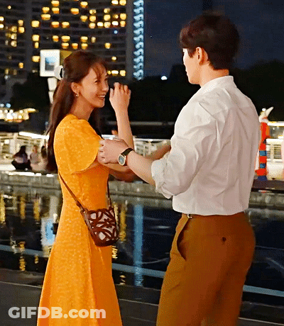 A Woman In A Yellow Dress Holds A Man 'S Hand In Front Of A Body Of Water GIF