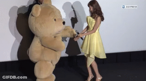 A Woman In A Yellow Dress Holds A Microphone Next To A Teddy Bear Costume GIF