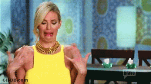 A Woman In A Yellow Dress Is Sitting At A Table With Her Arms Outstretched And Making A Funny Face . GIF