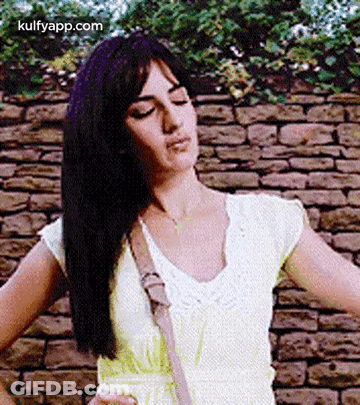 A Woman In A Yellow Dress Is Standing In Front Of A Brick Wall With Her Hands On Her Hips . GIF