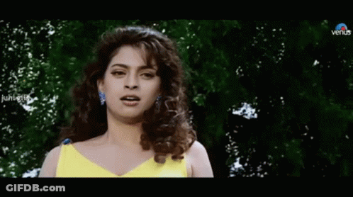 A Woman In A Yellow Dress Is Standing In Front Of Trees And Looking At The Camera . GIF