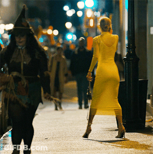 A Woman In A Yellow Dress Is Walking Down A Street At Night GIF