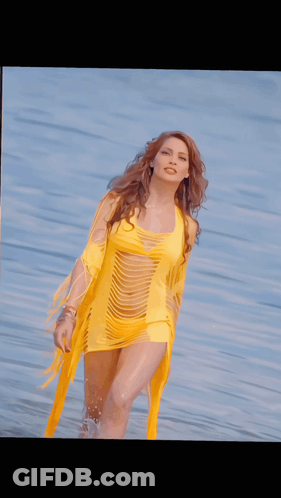 A Woman In A Yellow Dress Stands In Front Of A Body Of Water GIF