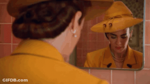 A Woman In A Yellow Hat Is Looking At Herself In A Mirror . GIF