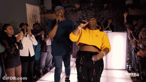 A Woman In A Yellow Hoodie Sings Into A Microphone In Front Of A Crowd GIF