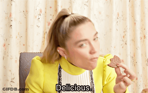 A Woman In A Yellow Jacket Is Eating A Cookie And The Words Delicious Are Above Her GIF