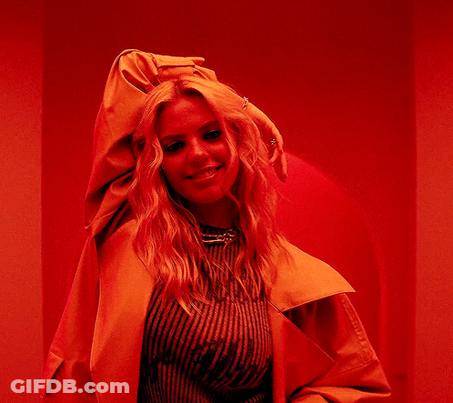 A Woman In A Yellow Jacket Is Standing In Front Of A Red Wall And Smiling . GIF
