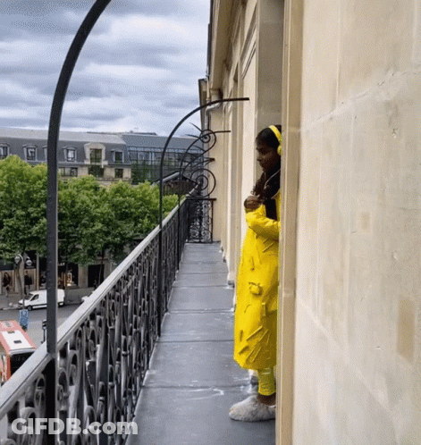 A Woman In A Yellow Raincoat Looks Out From A Balcony GIF