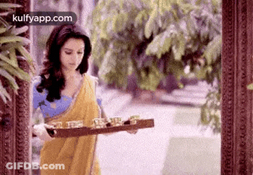 A Woman In A Yellow Sari Is Holding A Wooden Tray With Glasses On It . GIF