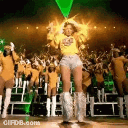 A Woman In A Yellow Shirt And Shorts Is Dancing On A Stage In Front Of A Crowd Of People . GIF
