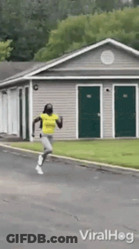 A Woman In A Yellow Shirt Is Running Down A Street In Front Of A House . GIF