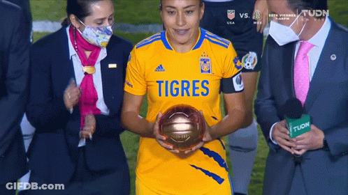 A Woman In A Yellow Shirt That Says Tigres Is Holding A Gold Ball GIF