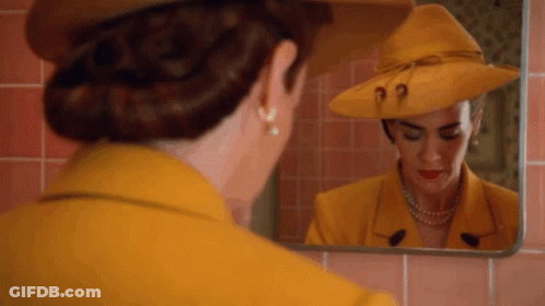A Woman In A Yellow Suit And Hat Is Looking At Herself In A Mirror . GIF