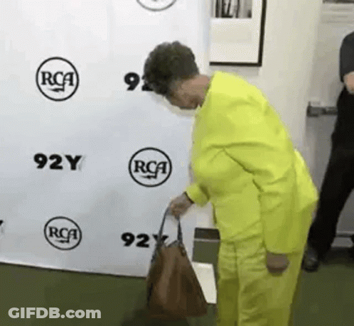 A Woman In A Yellow Suit Is Holding A Brown Purse In Front Of A Rca Sign . GIF