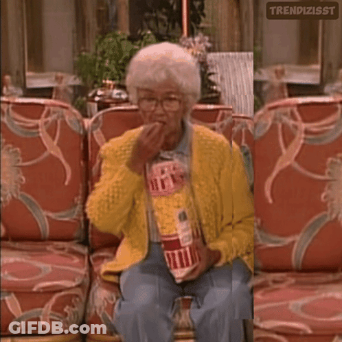 A Woman In A Yellow Sweater Is Sitting On A Couch Holding A Bag Of Popcorn GIF