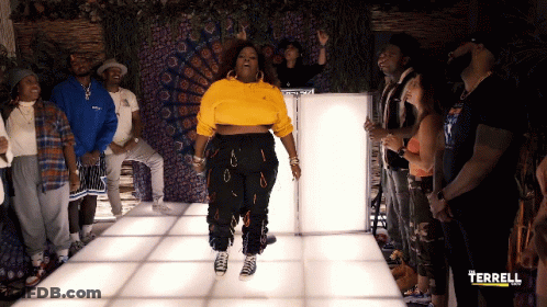 A Woman In A Yellow Sweatshirt Is Standing In Front Of A Group Of People With The Word Terrell On The Bottom Left GIF