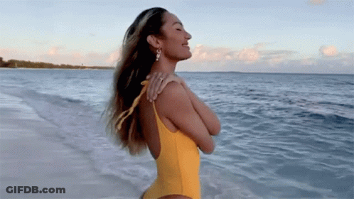 A Woman In A Yellow Swimsuit Is Standing On The Beach Looking At The Ocean . GIF