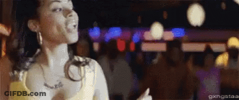 A Woman In A Yellow Tank Top Is Dancing In A Crowded Room . GIF