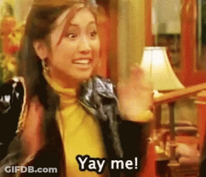 A Woman In A Yellow Turtleneck And Black Jacket Is Waving Her Hand And Saying Yay Me . GIF
