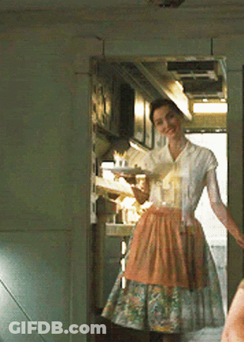 A Woman In An Apron Is Standing In A Kitchen Holding A Tray GIF