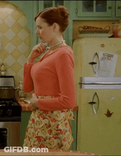 A Woman In An Apron Is Standing In A Kitchen With A Yellow Refrigerator GIF