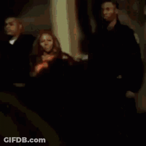 A Woman In An Orange Dress Is Standing Between Two Men In Black Suits GIF