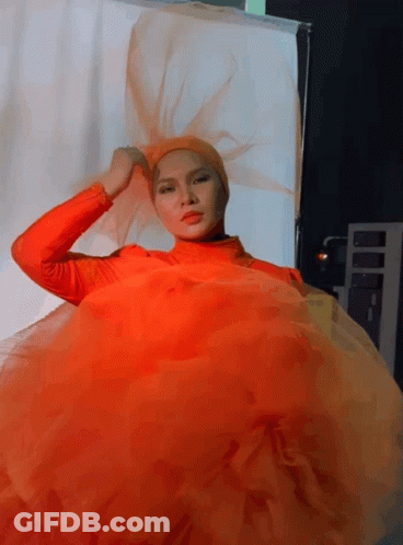 A Woman In An Orange Dress Is Standing In Front Of A White Backdrop GIF