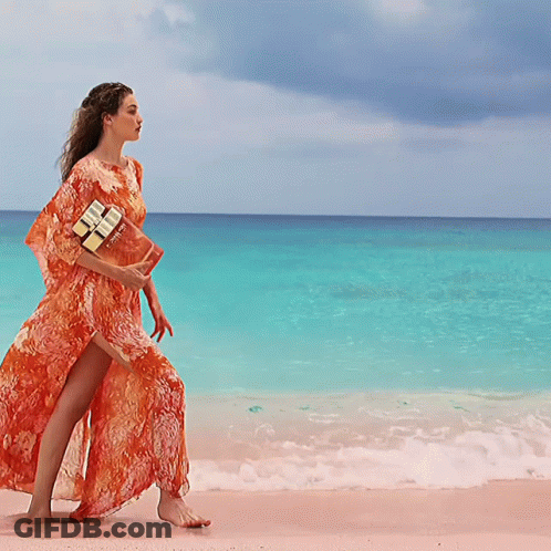 A Woman In An Orange Dress Walking On The Beach GIF