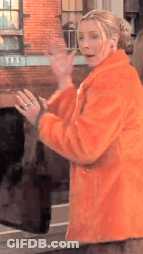 A Woman In An Orange Fur Coat Is Standing In Front Of A Brick Building And Waving . GIF