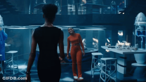A Woman In An Orange Jumpsuit Stands Next To Another Woman In A Black Dress GIF