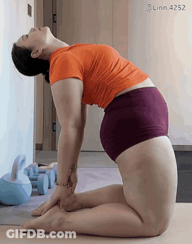A Woman In An Orange Shirt And Purple Shorts Is Doing Yoga On A Mat GIF