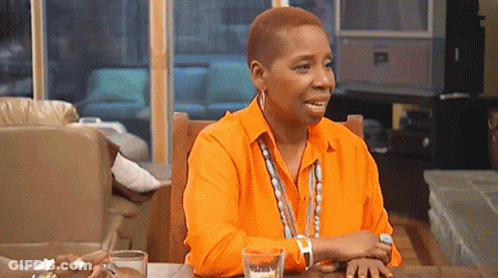 A Woman In An Orange Shirt Is Sitting At A Table With A Glass Of Water And A Pen . GIF