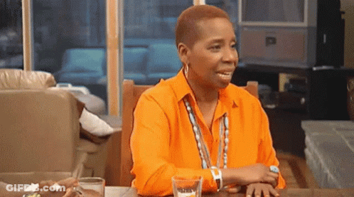 A Woman In An Orange Shirt Is Sitting At A Table With A Glass Of Water And A Watch . GIF