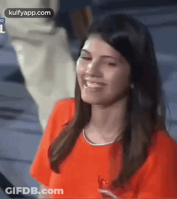 A Woman In An Orange Shirt Is Smiling And Looking At The Camera While A Man Stands Behind Her . GIF