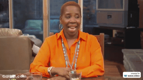 A Woman In An Orange Shirt Sits At A Table With Two Glasses Of Water GIF