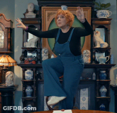 A Woman In Blue Overalls Stands On One Leg In Front Of A Fireplace GIF