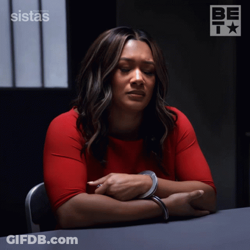 A Woman In Handcuffs Sits At A Table In Front Of A Sign That Says Sistas On It GIF