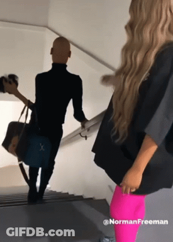 A Woman In Neon Pink Leggings Is Standing Next To A Man In A Black Jacket GIF
