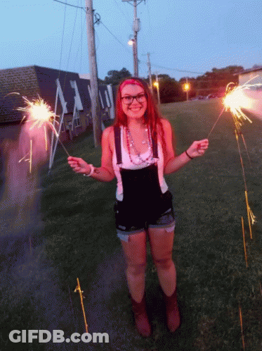 A Woman In Overalls Is Holding Sparklers In Front Of A Building That Says ' A ' On It GIF