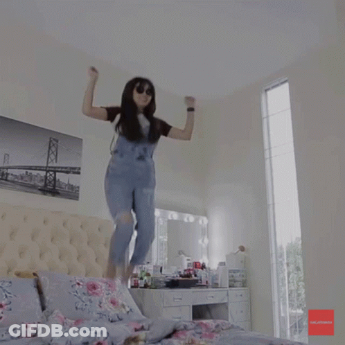 A Woman In Overalls Is Jumping In The Air In A Bedroom With A Picture Of A Bridge On The Wall GIF