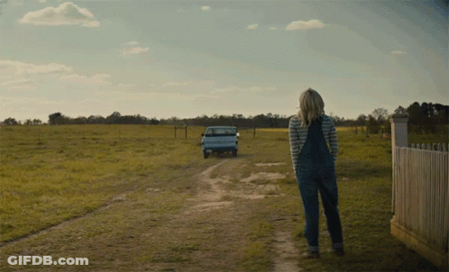 A Woman In Overalls Is Walking Down A Dirt Road With A Truck Behind Her GIF