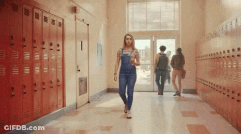 A Woman In Overalls Is Walking Down A Hallway In A School . GIF