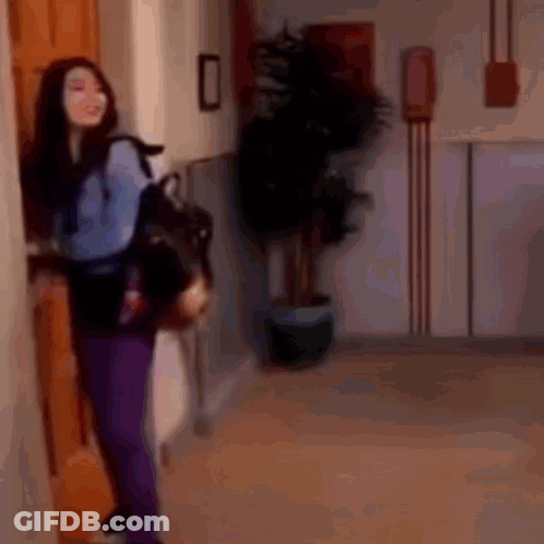 A Woman In Purple Pants Is Standing In A Hallway Holding A Backpack . GIF