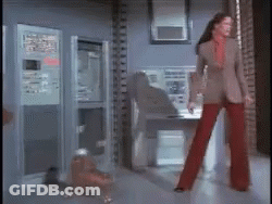 A Woman In Red Pants And A Brown Jacket Is Standing In A Room GIF