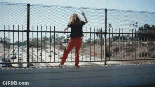 A Woman In Red Pants Is Standing On A Bridge Looking Over A Fence GIF