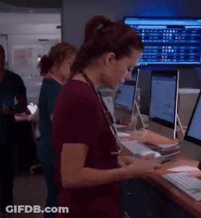 A Woman In Scrubs Is Looking At A Tablet In Front Of A Computer Monitor . GIF