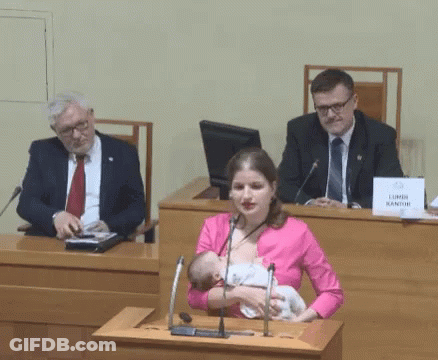 A Woman Is Breastfeeding A Baby In Front Of A Podium With A Sign That Says Lumen Kantor GIF