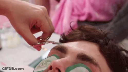 A Woman Is Cutting A Man 'S Eyebrow With Scissors While He Wears Cucumber Slices On His Eyes . GIF