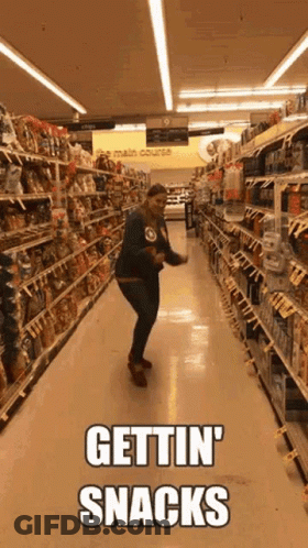 A Woman Is Dancing In A Grocery Store Aisle With The Words Gettin ' Snacks Written On It . GIF
