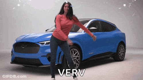 A Woman Is Dancing In Front Of A Blue Ford Mustang With The Word Very Above Her GIF